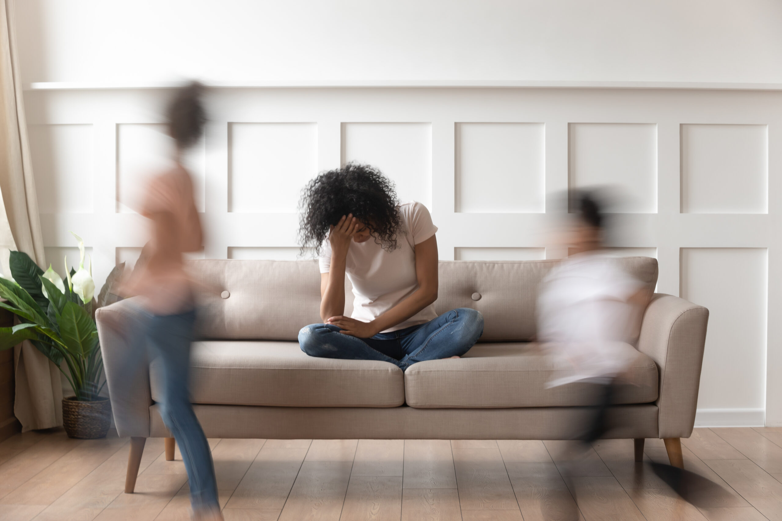 Co-Parenting Stress – Finding Calm Amid the Chaos A stressed mother sits on a couch holding her head while two children run around her in a blurred motion, symbolizing the overwhelm and chaos often felt in co-parenting situations.
