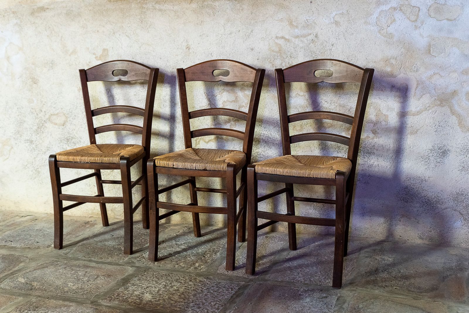 Three wooden chairs side by side against a sunlit wall, for the lawyer, the therapist and the divorce coach